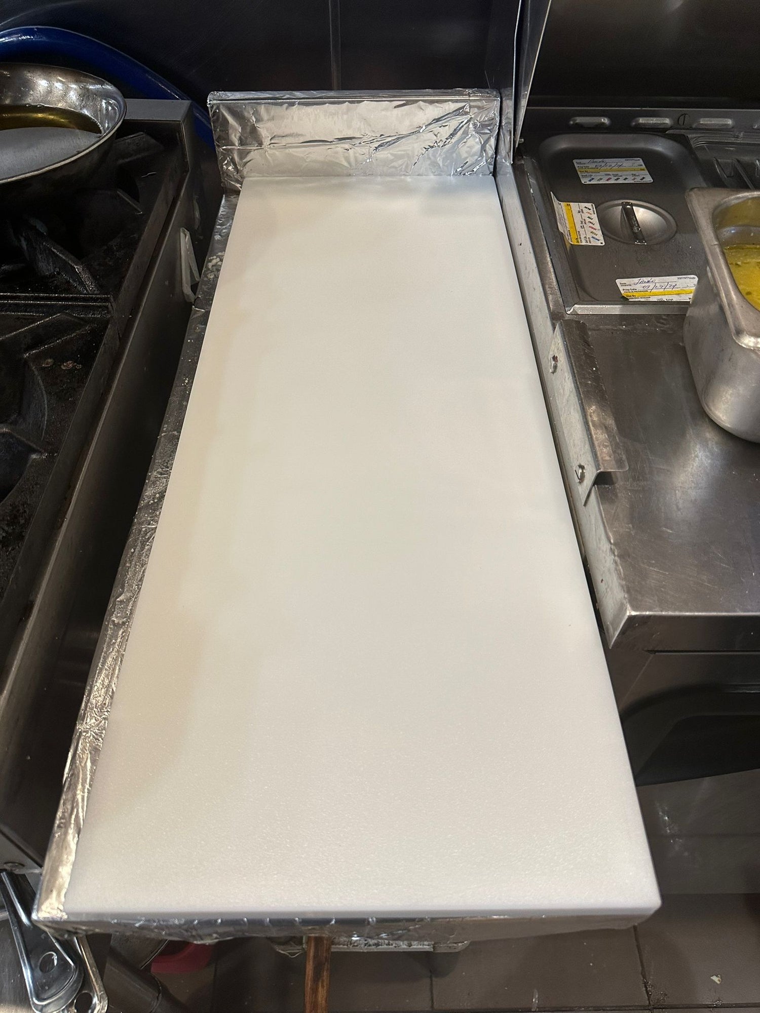 Clean cutting board placed in a professional kitchen environment, surrounded by cooking equipment and utensils.